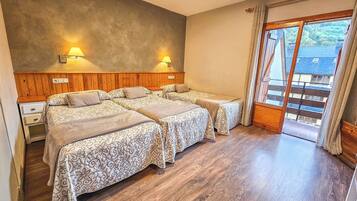 Una camera d'albergo con pavimento in legno, due letti con copriletto decorato, un comodino e una finestra che si affaccia su edifici.