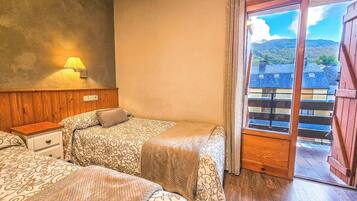 A room with two beds, a nightstand, a lamp, and a view of buildings and mountains through a window.