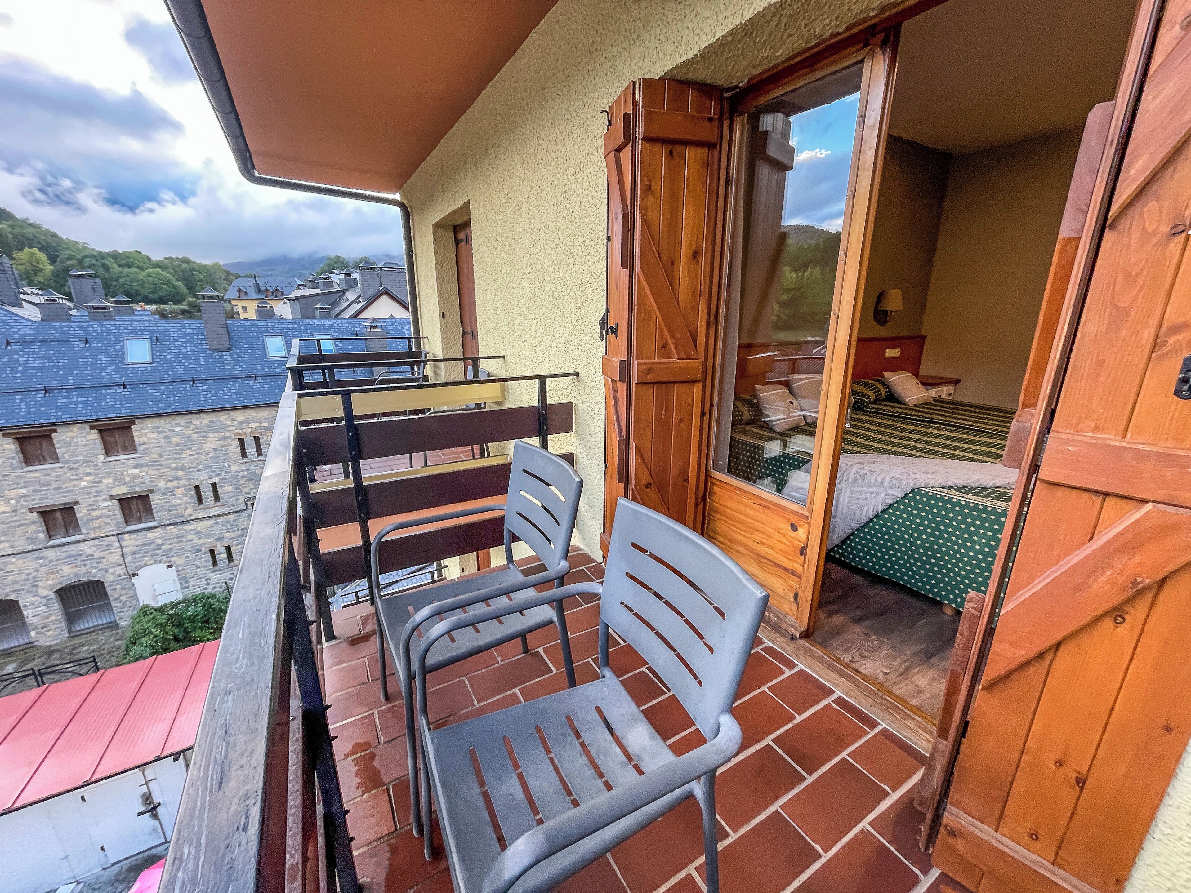 Een balkon met twee metalen stoelen en een houten deur naar een kamer met een bed.