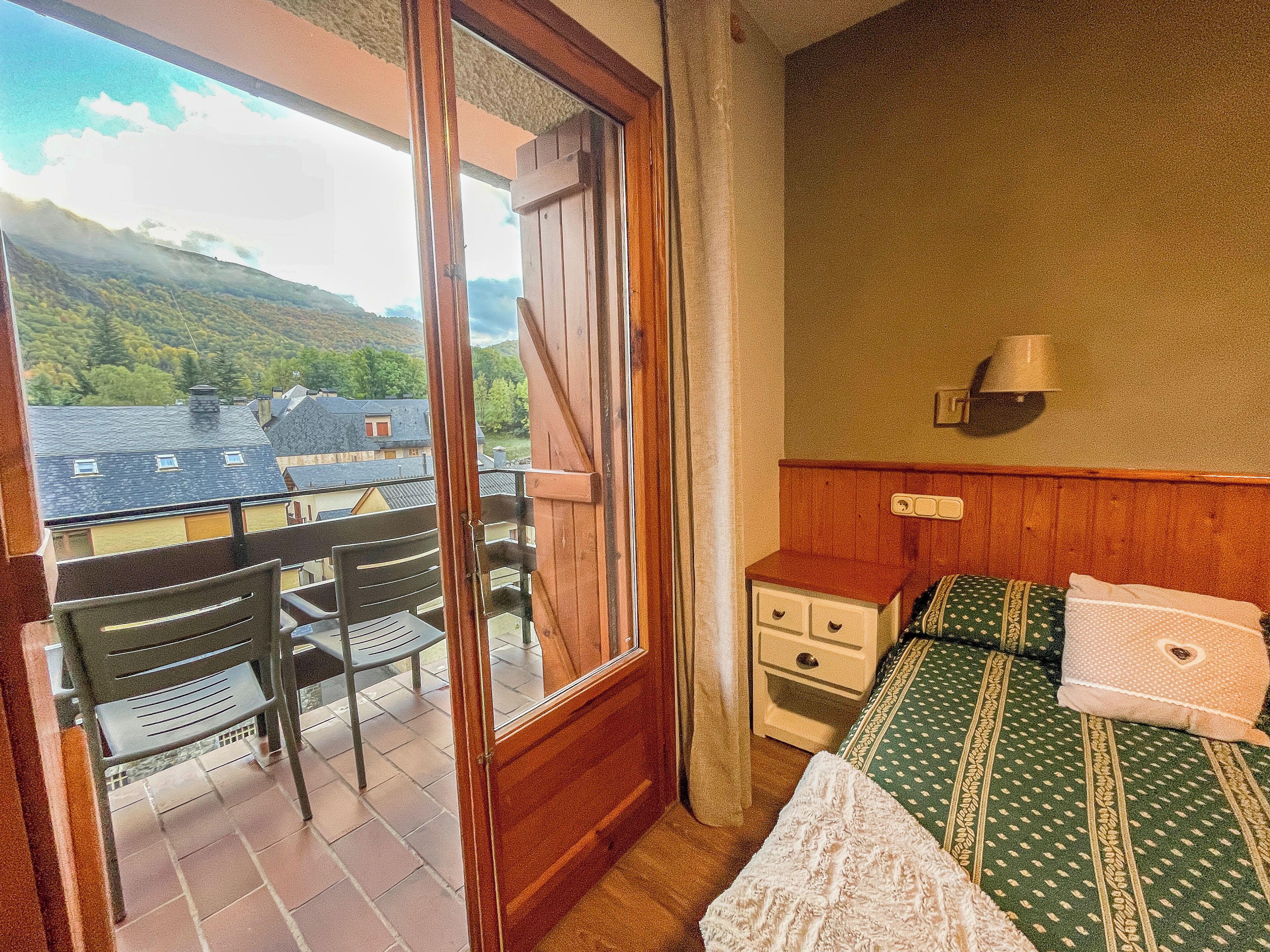 A room with a balcony, a bed with patterned bedding, a nightstand, and a wall-mounted lamp.