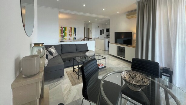 A modern living room with a sofa, coffee table, and chairs, a TV, and a kitchen area in the background. - 2-bed Sea View Apt, 10 Mins From Puerto Banus (Marbella)