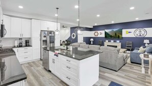 Une cuisine moderne avec des armoires blanches, un comptoir en granit et des appareils électroménagers en acier inoxydable. On y trouve un coin repas avec un canapé gris et une petite table. Le mur d’accent est bleu et est agrémenté d’objets décoratifs et d’une horloge.