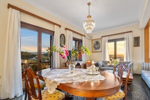 Un coin repas avec une table ronde en bois, une nappe en dentelle et un lustre. Des chaises sont disposées autour de la table, et un canapé se trouve au fond de la pièce.