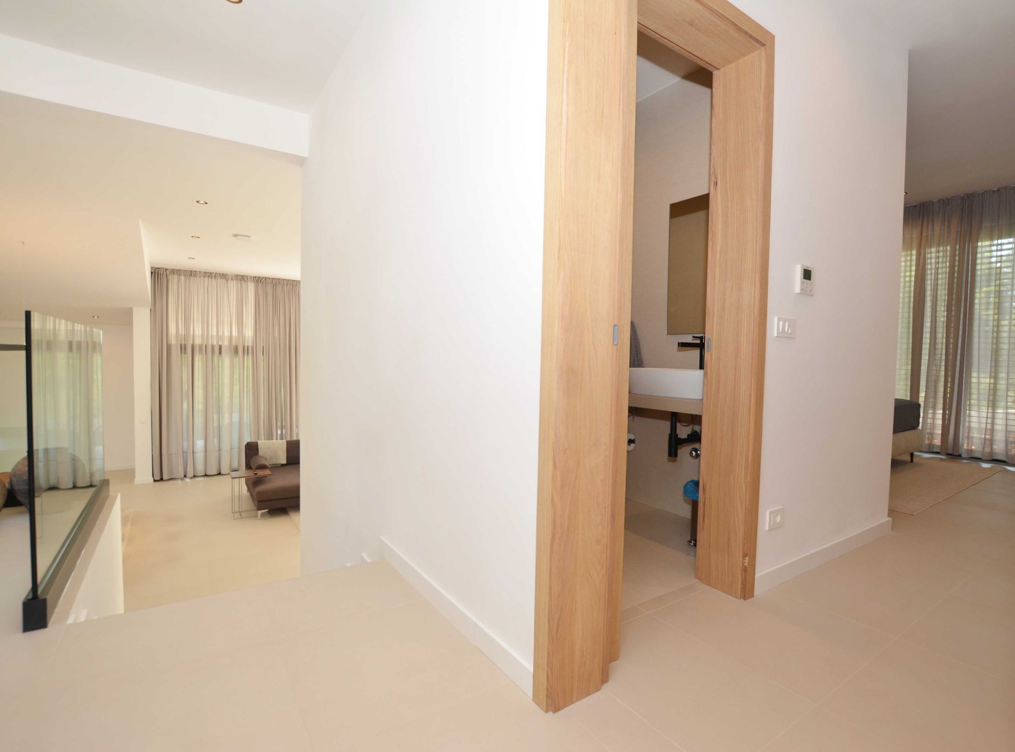 Een moderne hotelkamer met een houten deur naar een badkamer, een bank in de zithoek en grote ramen met gordijnen.