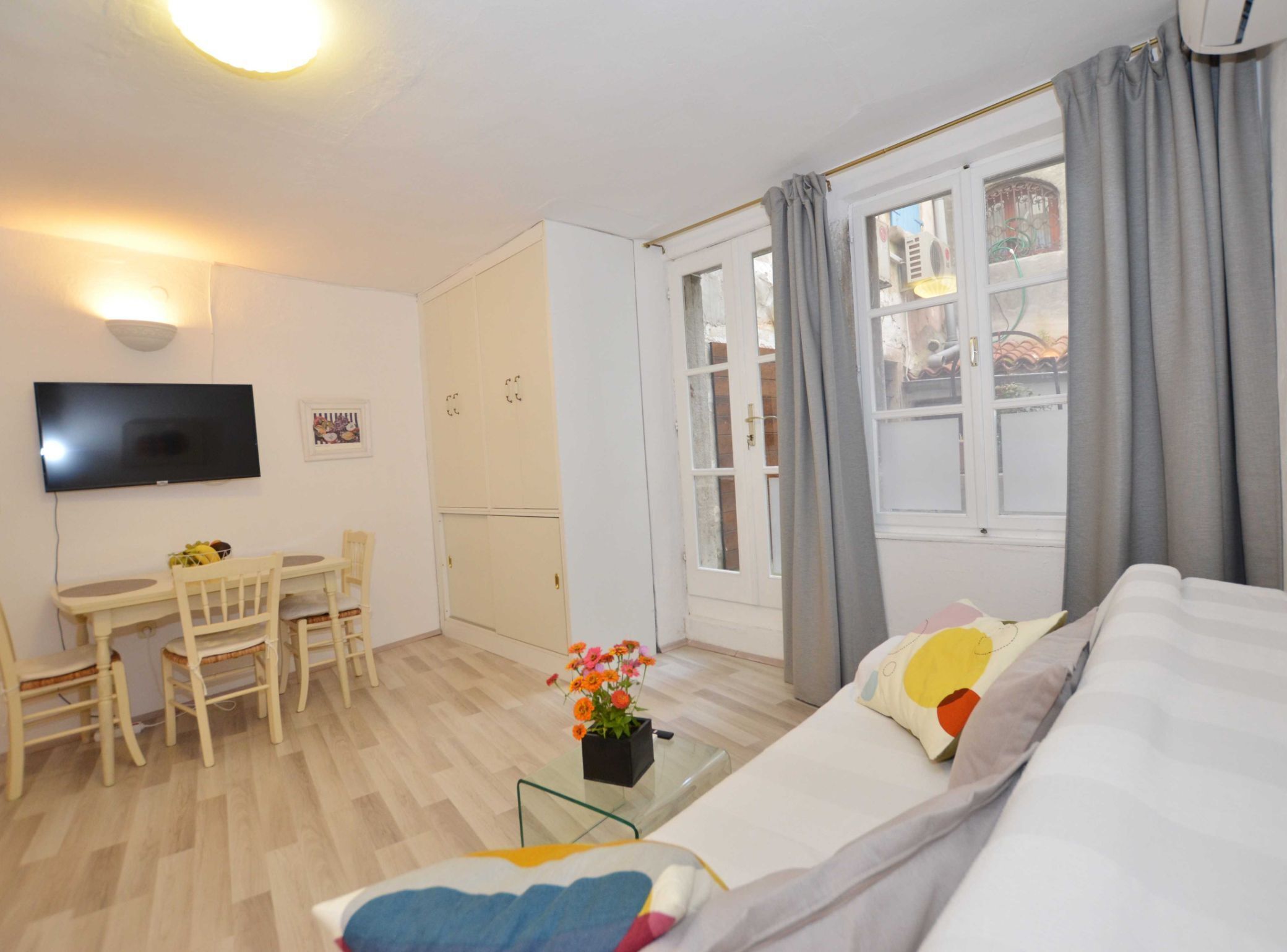 A hotel room with a bed, a TV, a desk with chairs, a window with curtains, and a small table with a flower vase.