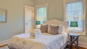 A neatly made bed with a stack of white towels, two bedside lamps, and a window with blinds.