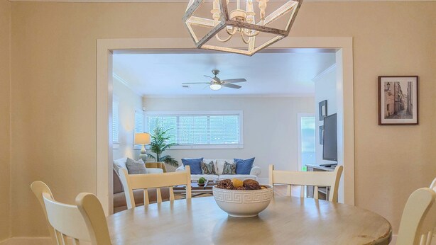 A dining area with a round table, white chairs, and a chandelier. There is a ceiling fan and a window with blinds in the background.