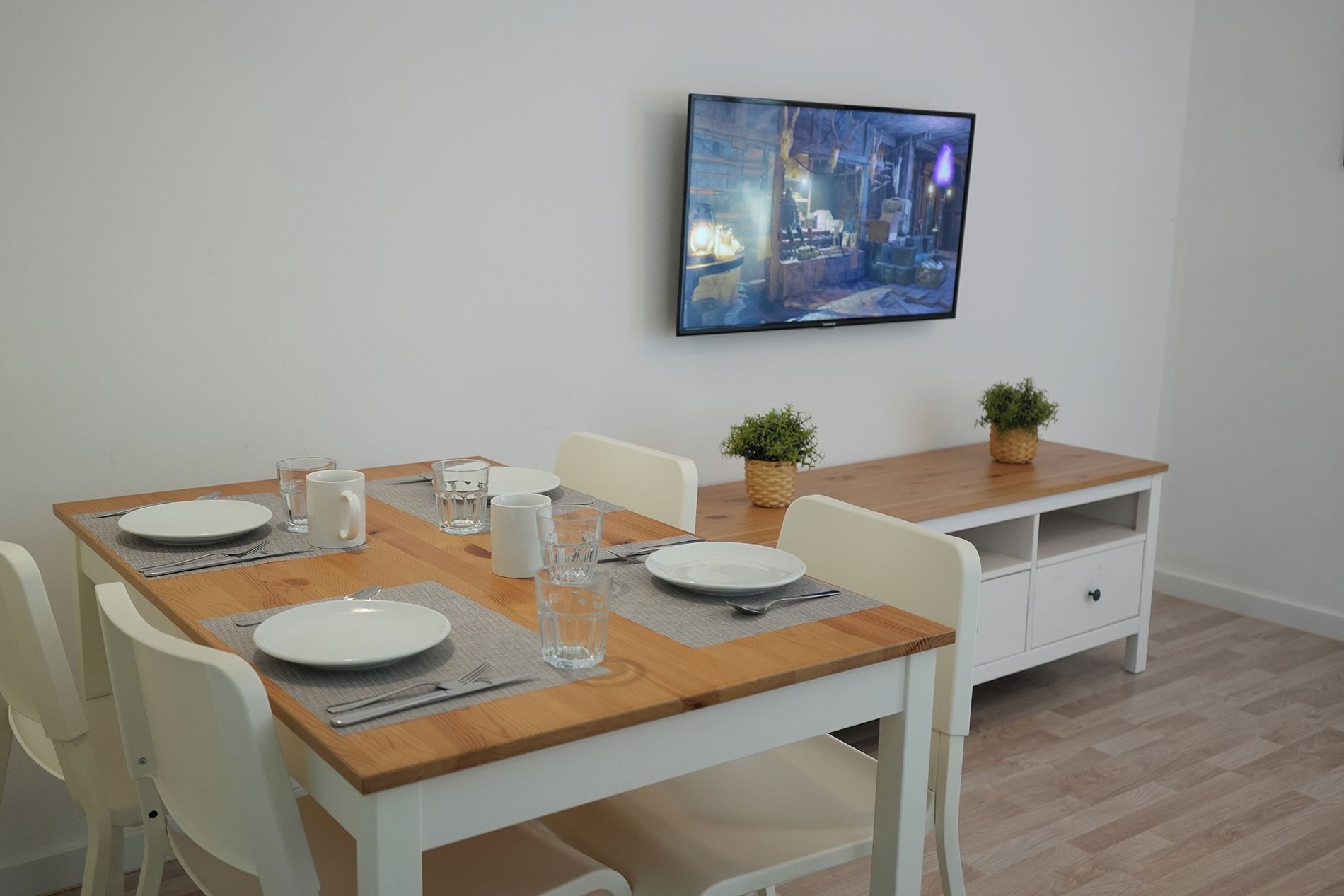 Een eetgedeelte met een houten tafel, witte stoelen en een aan de muur bevestigde televisie.