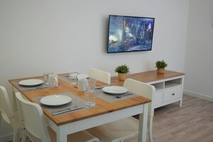 Un comedor con una mesa de madera, sillas blancas y un televisor montado en la pared.