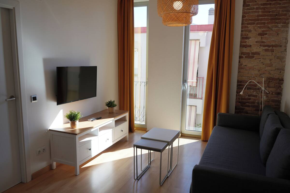 A modern living room with a flat-screen TV, a wooden desk, a grey sofa, and a balcony with orange curtains.