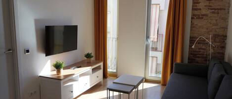 A modern living room with a flat-screen TV, a wooden desk, a grey sofa, and a balcony with orange curtains.