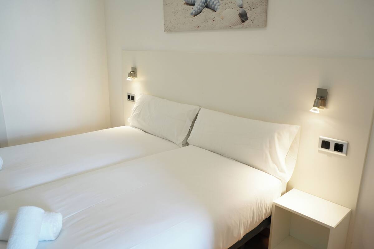 A large bed with white linens and pillows, two bedside tables with wall-mounted lamps, and a painting of sea creatures on the wall.