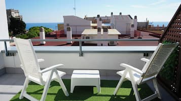 Un balcon avec deux chaises blanches et une petite table, d’où l’on peut admirer un paysage urbain avec ses bâtiments et un ciel dégagé.