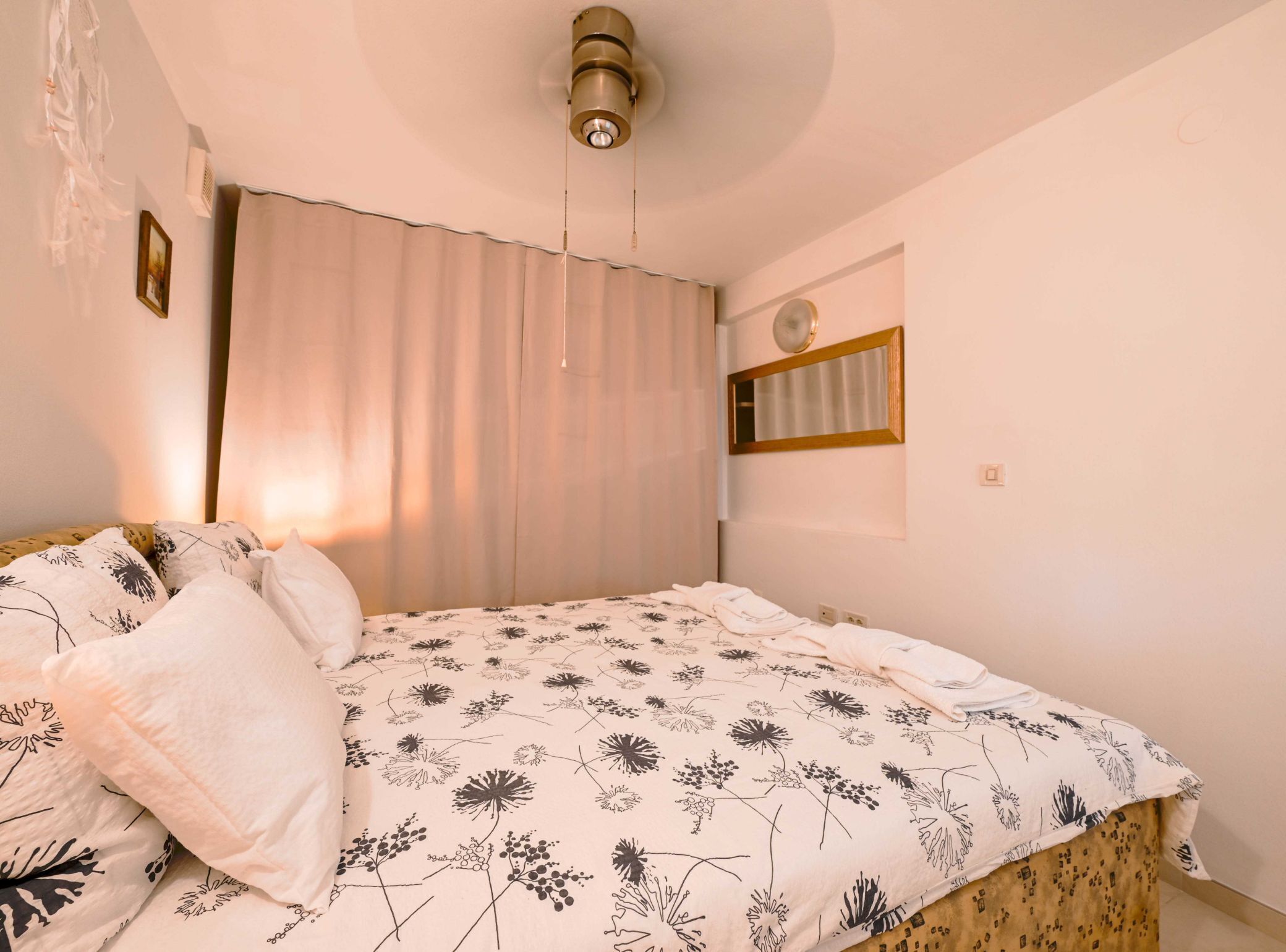 Un dormitorio con cama, ventana con cortinas, espejo montado en la pared y lámpara en el techo.
