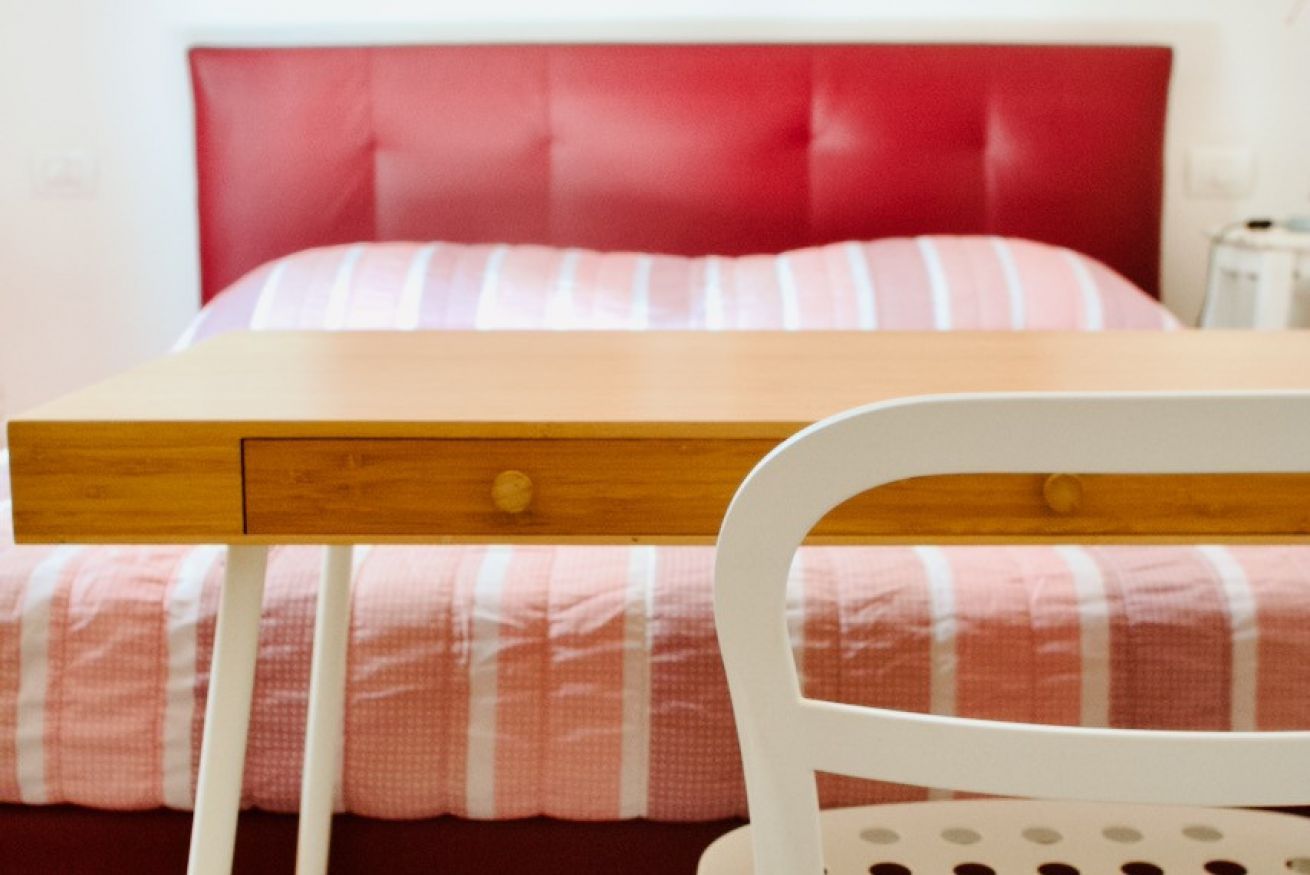 A bed with a red headboard, a wooden bedside table with drawers, and a white chair with a circular backrest.