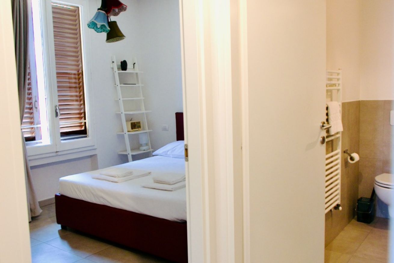 Ein Schlafzimmer mit Bett, Fenster mit Jalousien, Nachttisch, Ablagefläche und einem Badezimmer mit Toilette und Heizkörper.