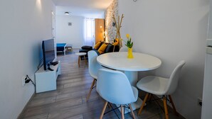 Ein modernes Wohnzimmer mit einem runden Esstisch, hellblauen Stühlen, einer Couch mit gelben Kissen und einem kleinen weißen Tisch mit einer gelben Vase.