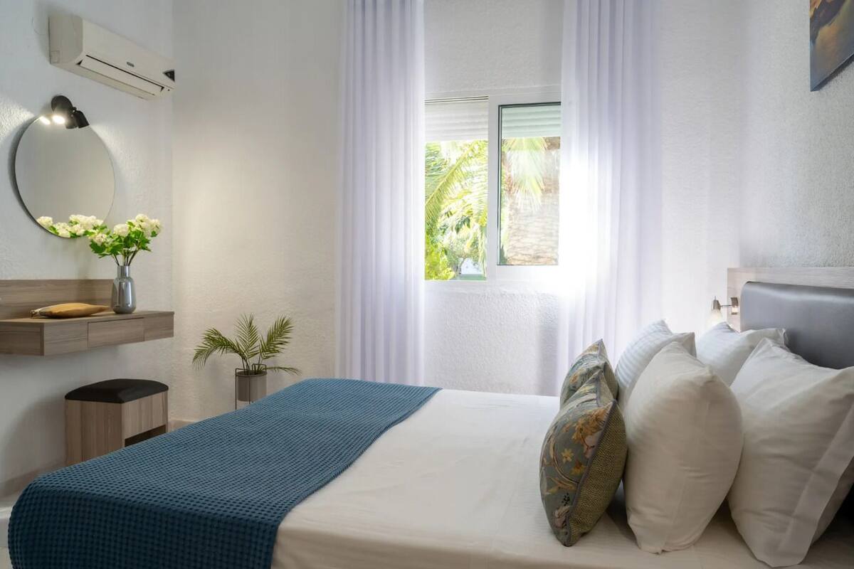 Une chambre d’hôtel comprenant un lit, une table de chevet, un miroir, une fenêtre avec des rideaux, une plante et un appareil de climatisation.