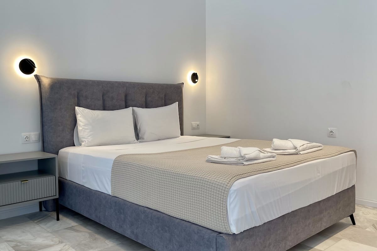 Une chambre à coucher moderne avec un lit recouvert d’un tissu gris, du linge de lit blanc, une table de chevet grise et deux lampes de lecture fixées au mur.