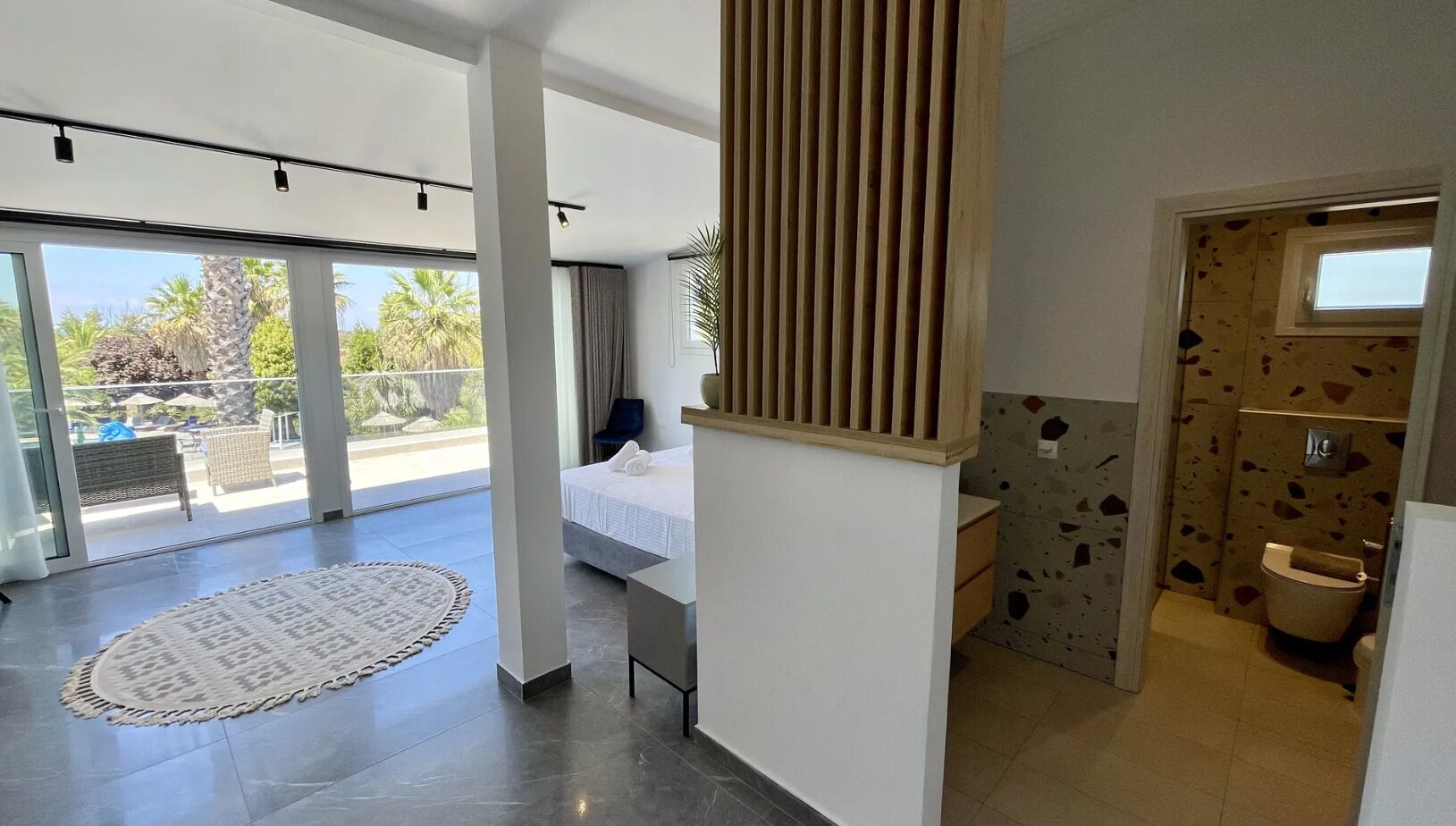 Ein modernes Hotelzimmer mit großem Fenster, einem Bett, einem Badezimmer mit einzigartigem Fleckenmuster und einem Balkon mit Gartenmöbeln.