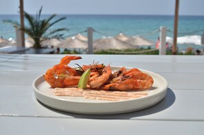 Une assiette de crevettes grillées est posée sur une table blanche, avec vue sur l’océan et des parasols de plage en arrière-plan.