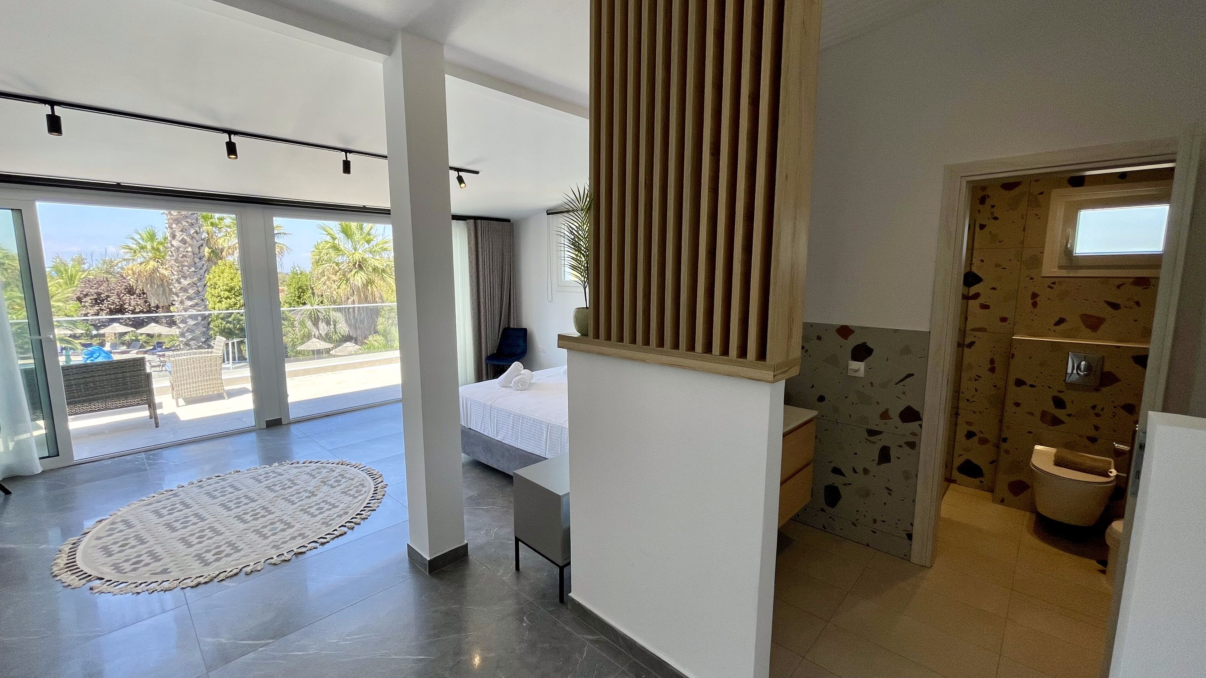 Una habitación de hotel moderna con balcón, un ventanal grande, una cama, un baño con un diseño de azulejos original y un panel de madera en la pared.