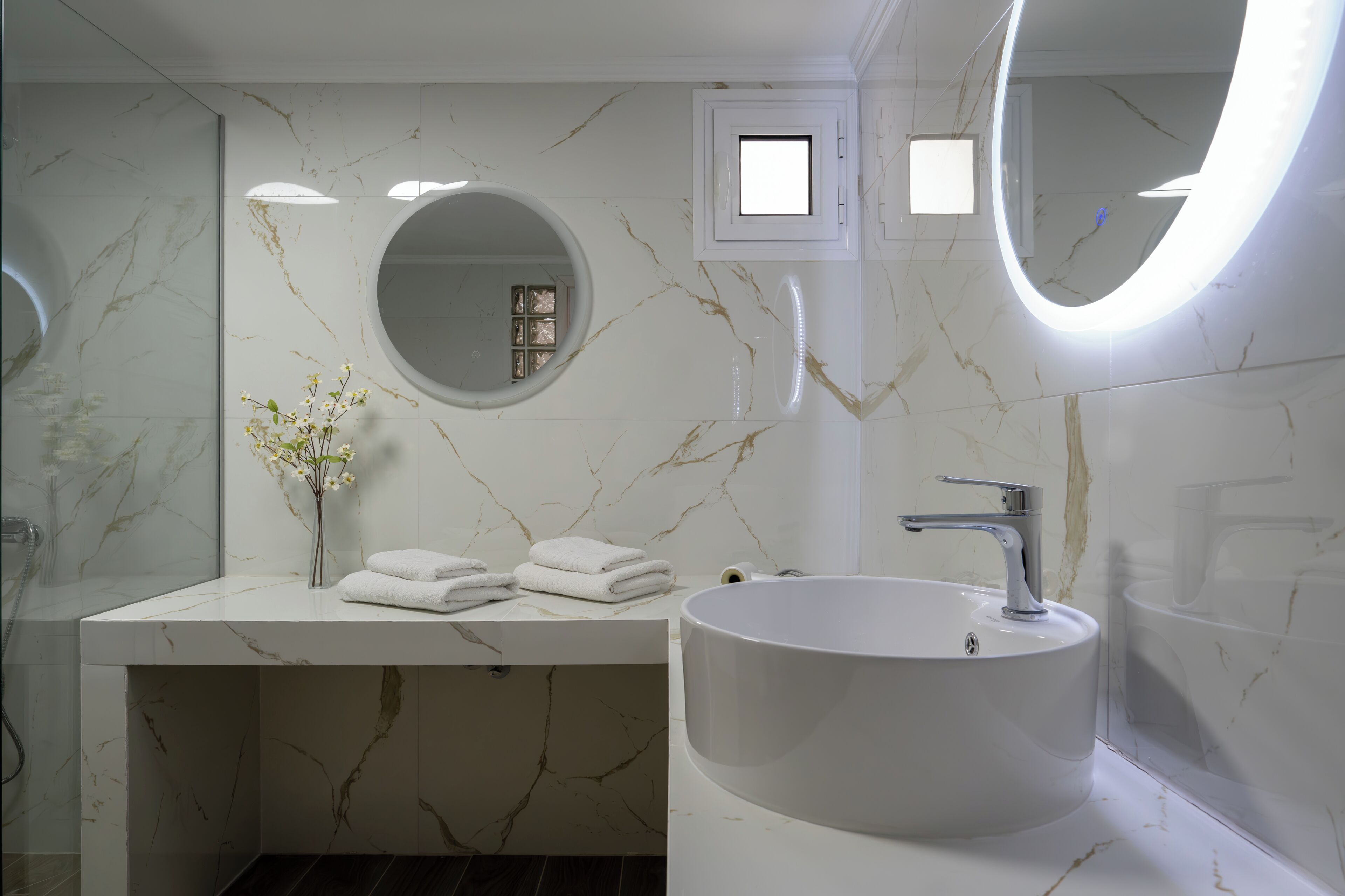 A bathroom with a marble vanity, a round mirror, a sink, and a vase with flowers.