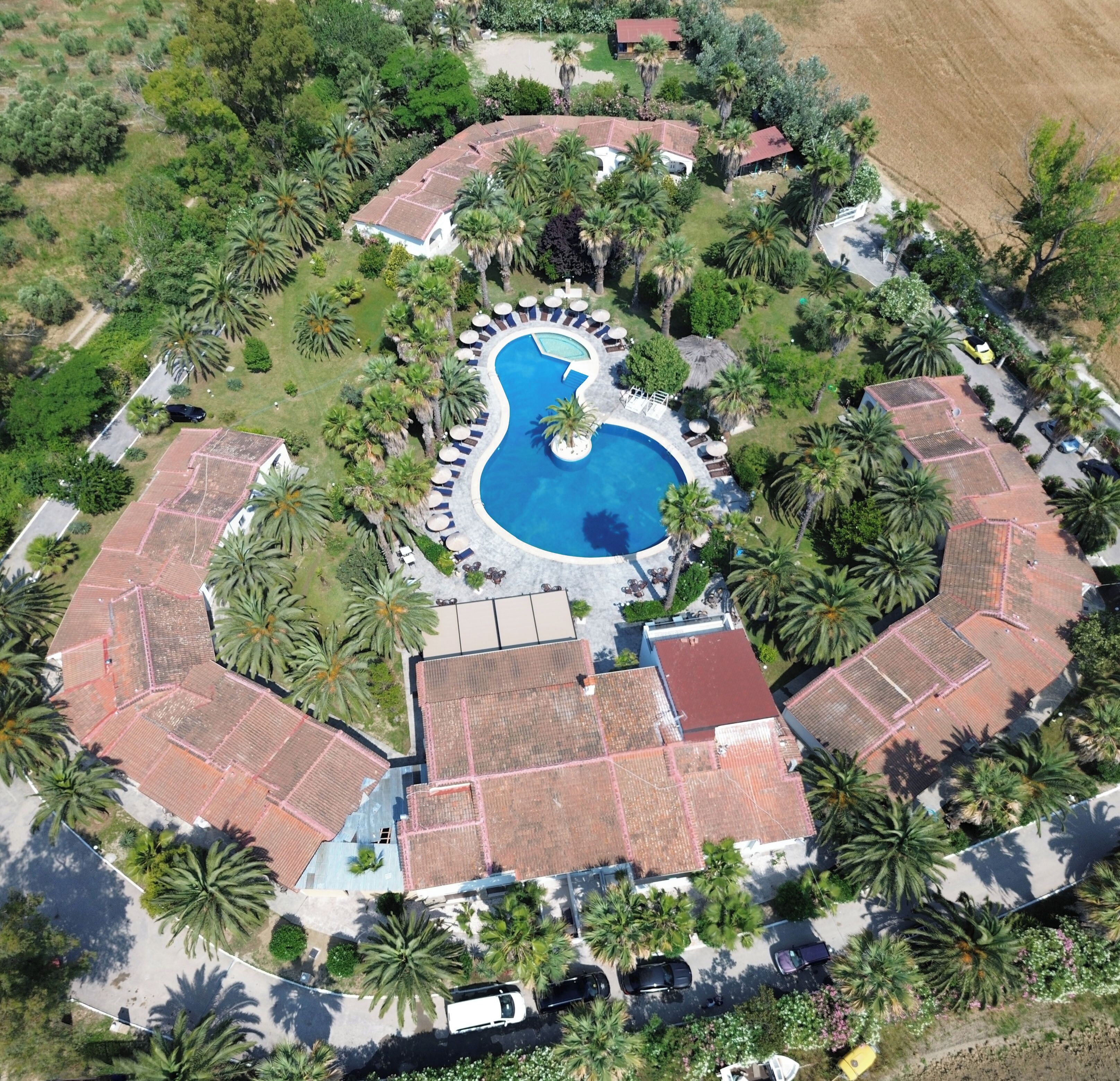 Vista aerea di un resort con una piscina centrale, circondata da palme e edifici con tetti in tegole rosse.