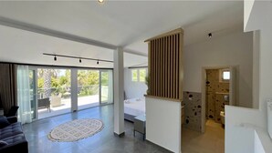 A spacious living area with a large sliding glass door leading to a balcony, a bed with white linens, and a modern kitchen area. - PALMA VILLAGE HOTEL by Greek Pride (Kassandra)