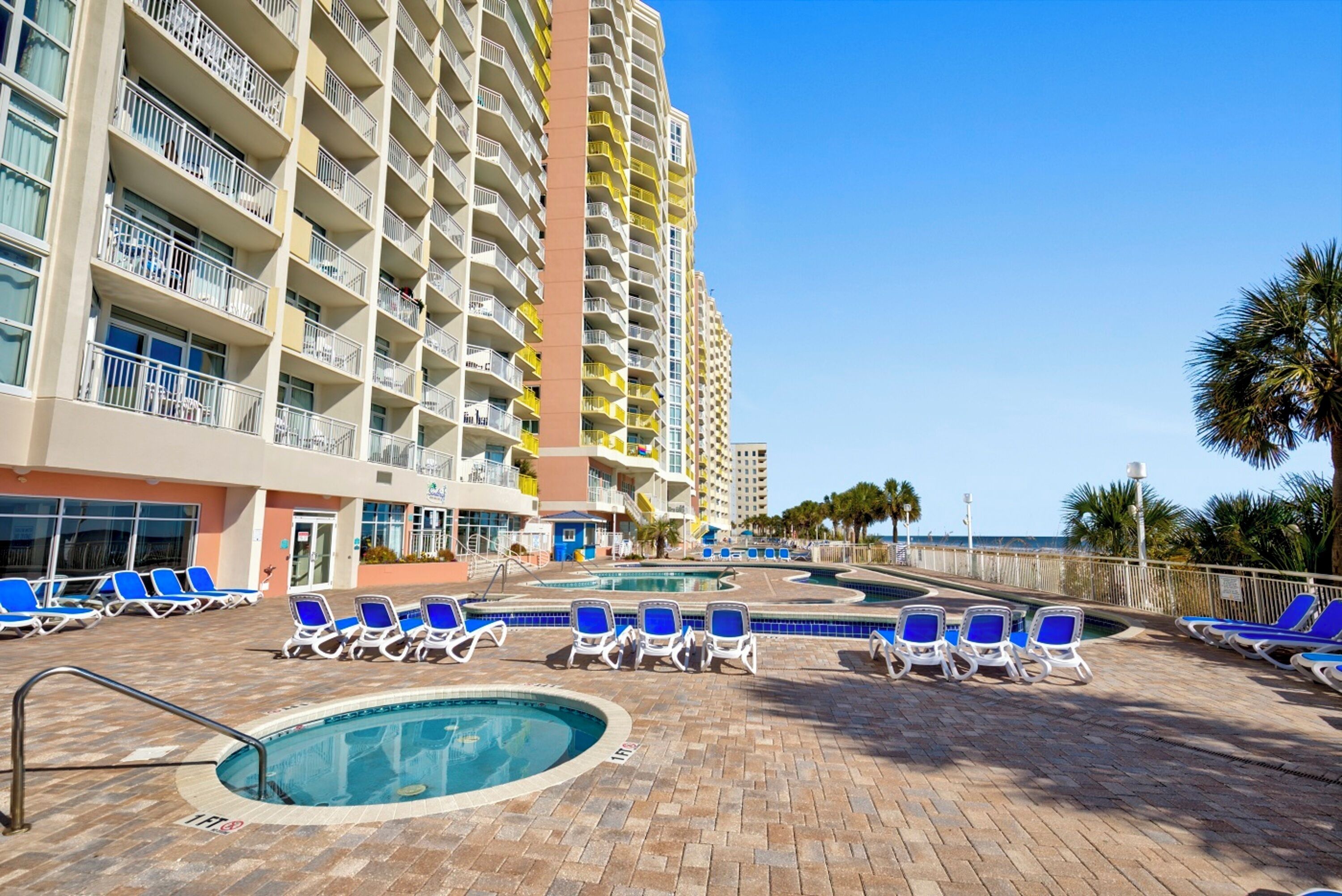 A hotel with multiple balconies, a swimming pool area with lounge chairs, and a view of the ocean.