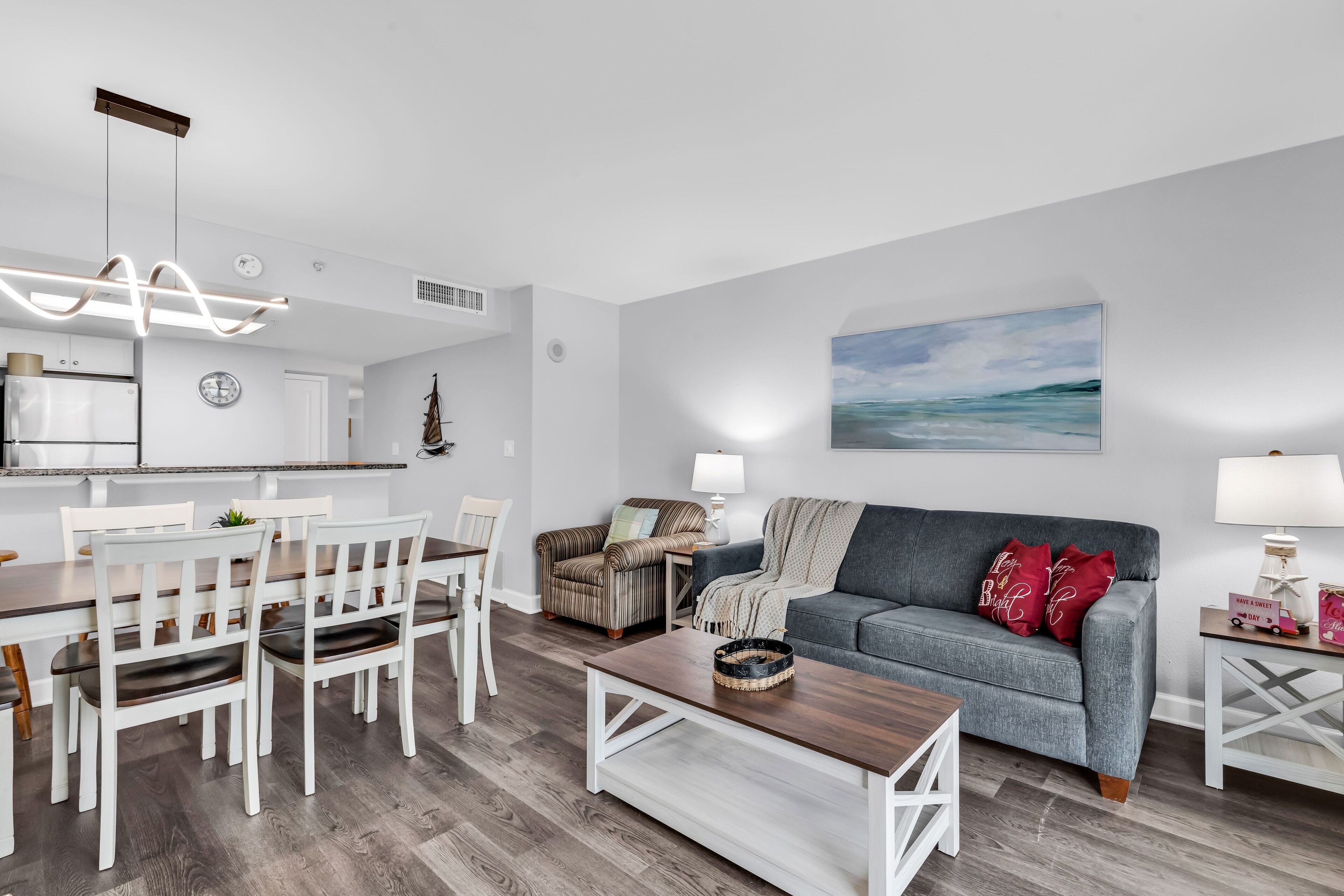 Een moderne woonkamer met een grijze bank, een witte eettafel en stoelen. Achtergrond is een keuken met witte kasten en een klok.