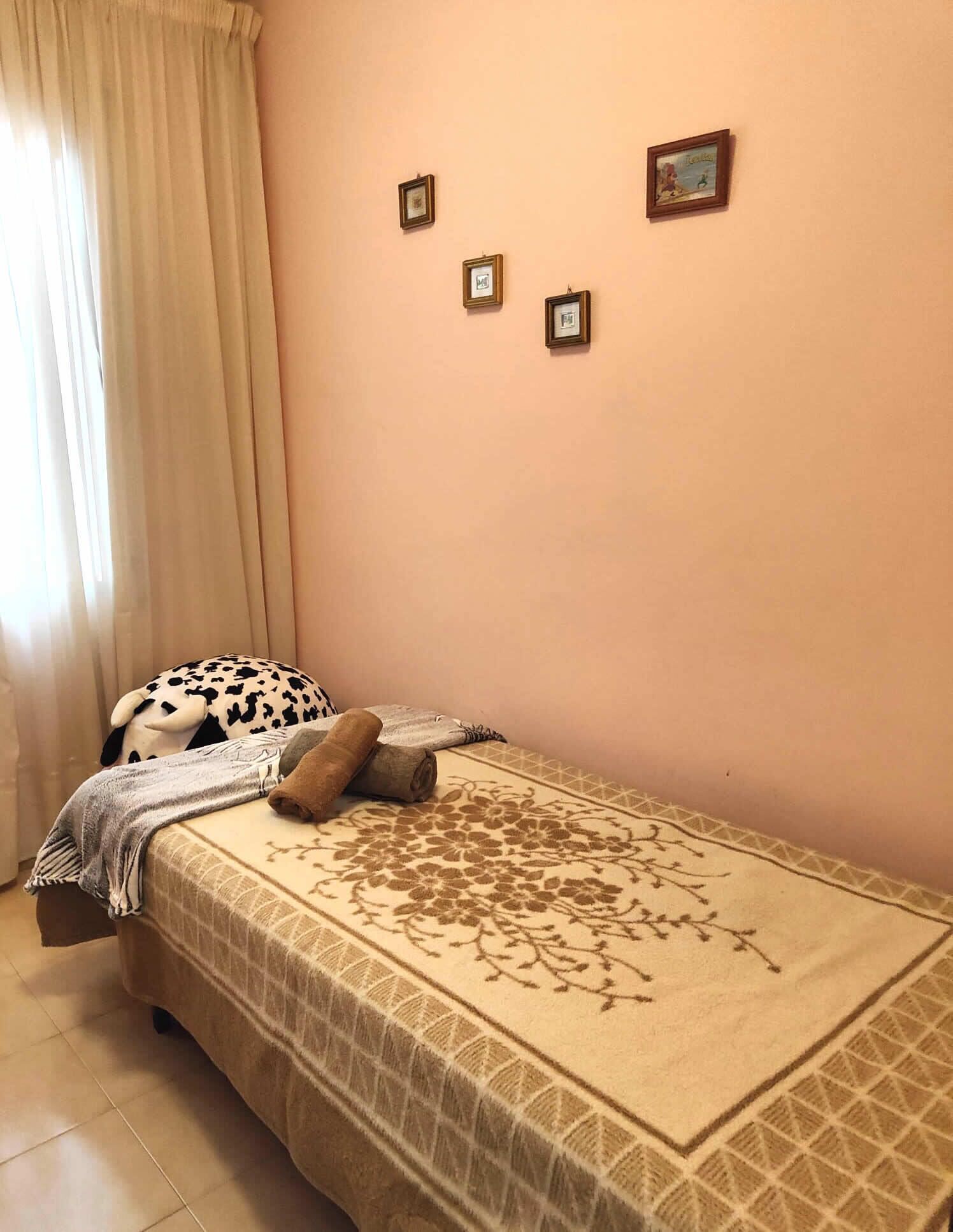 Un dormitorio pequeño y ordenado, con cama, ventana con cortinas y tres pequeños cuadros en la pared.