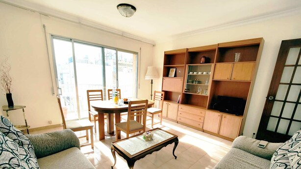 Una sala de estar con un juego de sofá, una mesa de centro, una mesa de comedor con sillas, un televisor sobre un mueble de madera y un ventanal con vistas a edificios.