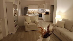 A modern living room with a sofa, a chair, and a coffee table. There is a kitchen in the background with white cabinets and a dining area.