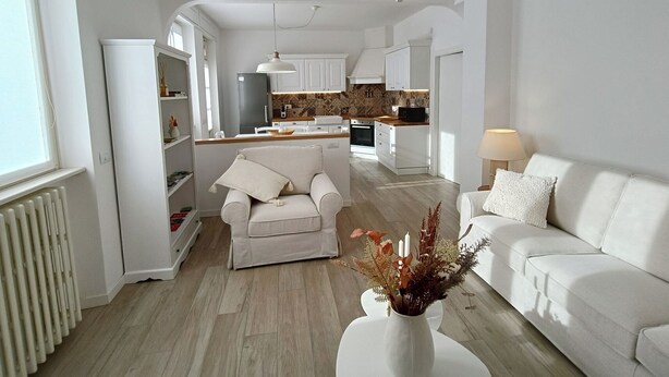 A modern living room with a sofa, a chair, a white vase with flowers, and a kitchen in the background.
