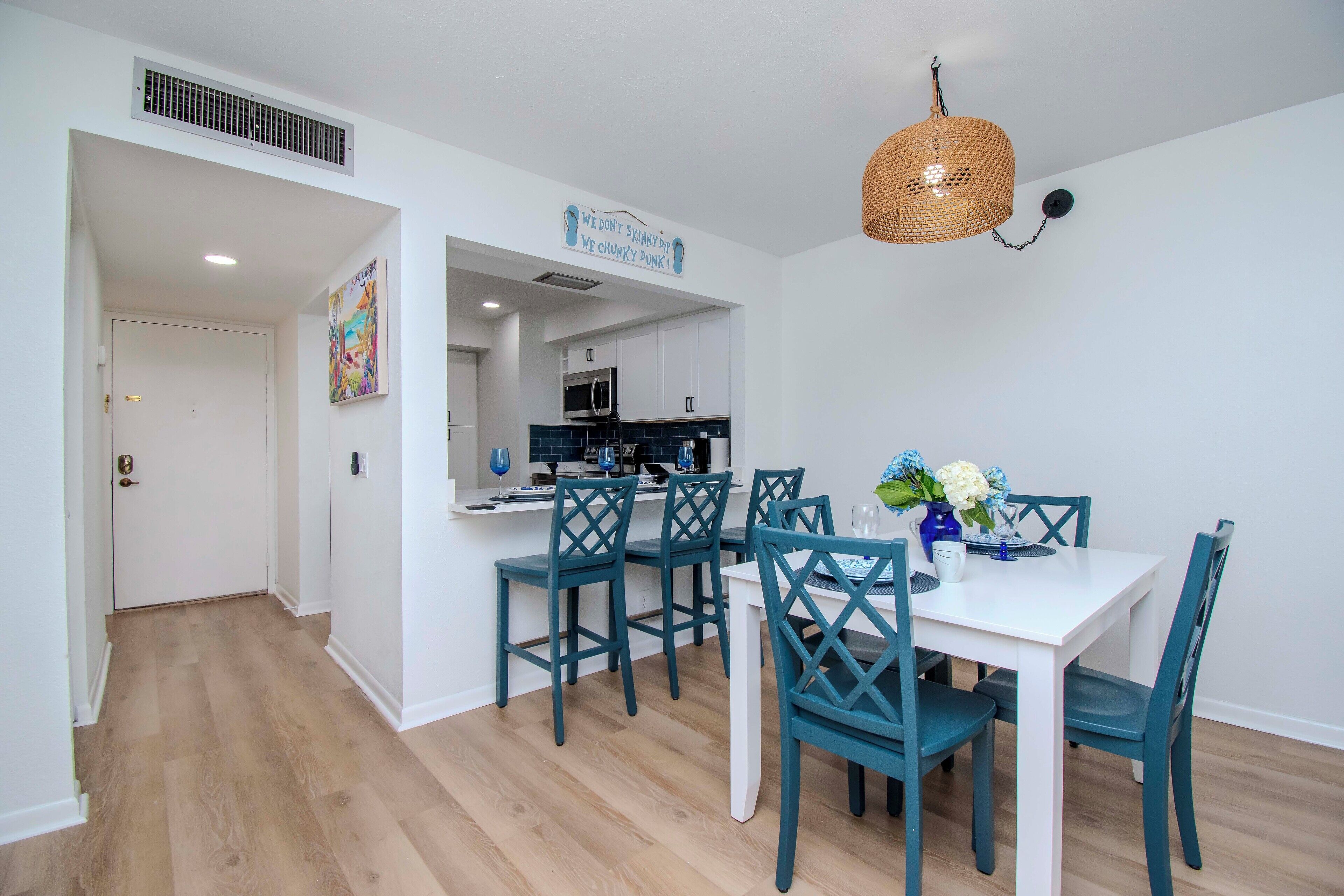 A modern kitchen with white cabinets, a microwave, and a sink. There is a dining area with a white table and teal chairs. A decorative sign hangs on the wall, and a woven light fixture is above the table.