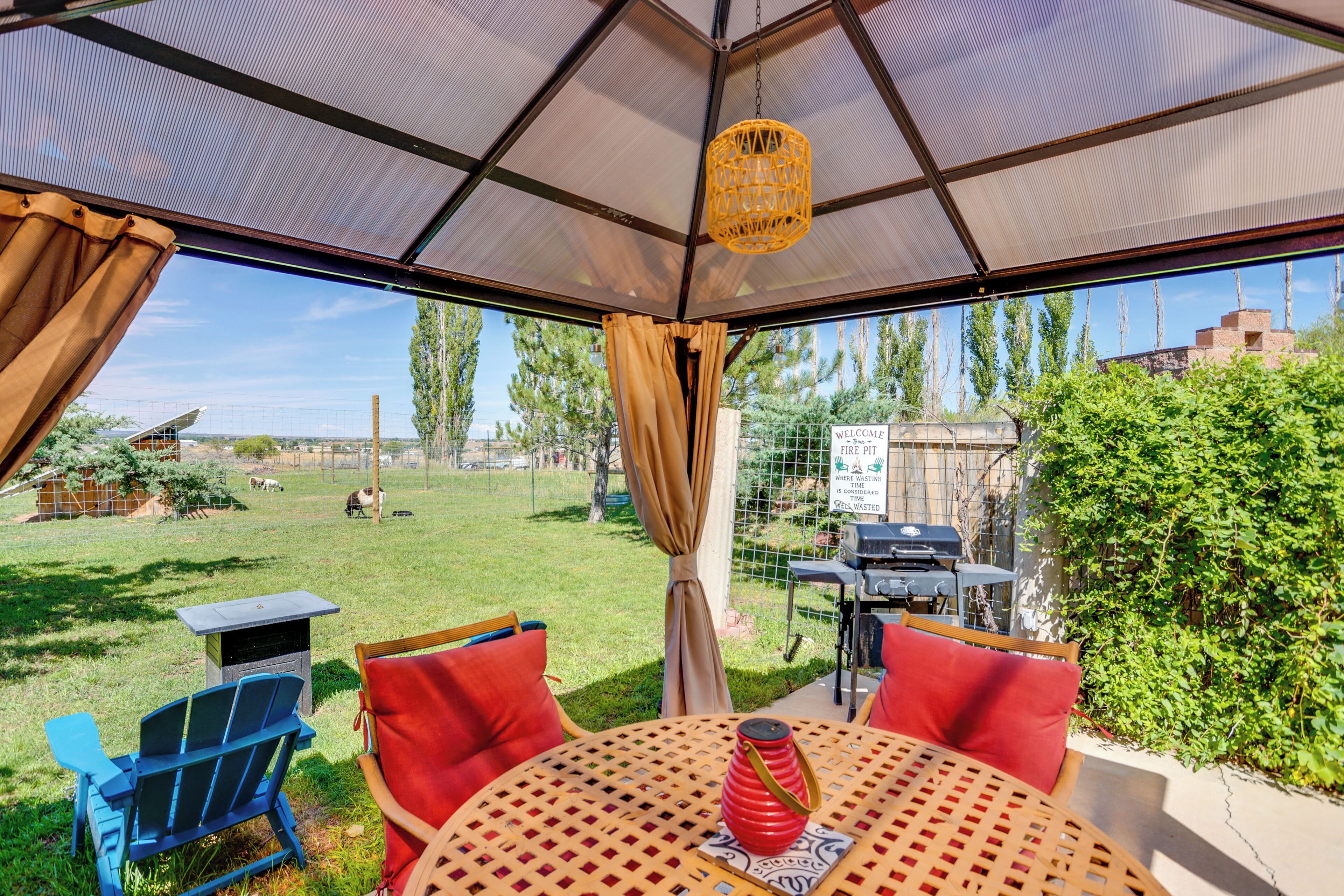 Eine Terrasse im Freien mit Tisch und Stühlen, einem Grill und Blick auf einen umzäunten Garten mit Bäumen und einem Gebäude in der Ferne.