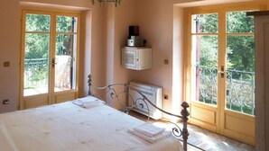 Ein Schlafzimmer mit einem großen Bett, Holzbalken, einer Chandelier, einem Heizkörper und zwei Fenstern mit Blick auf Grünflächen.