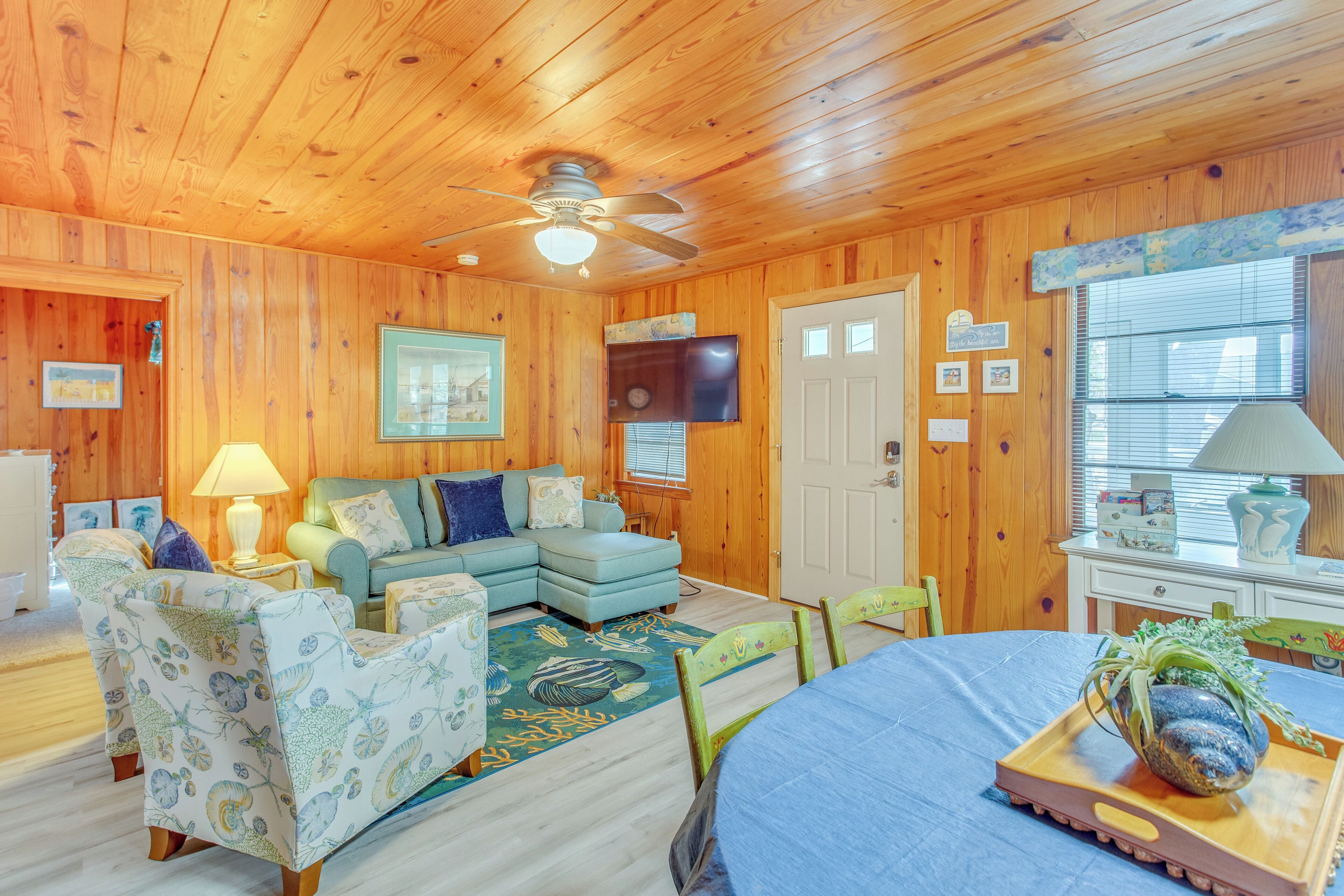 A cozy cabin interior with a wooden ceiling, a ceiling fan, a blue sofa, patterned chairs, a dining table with a tray, and a television on the wall.