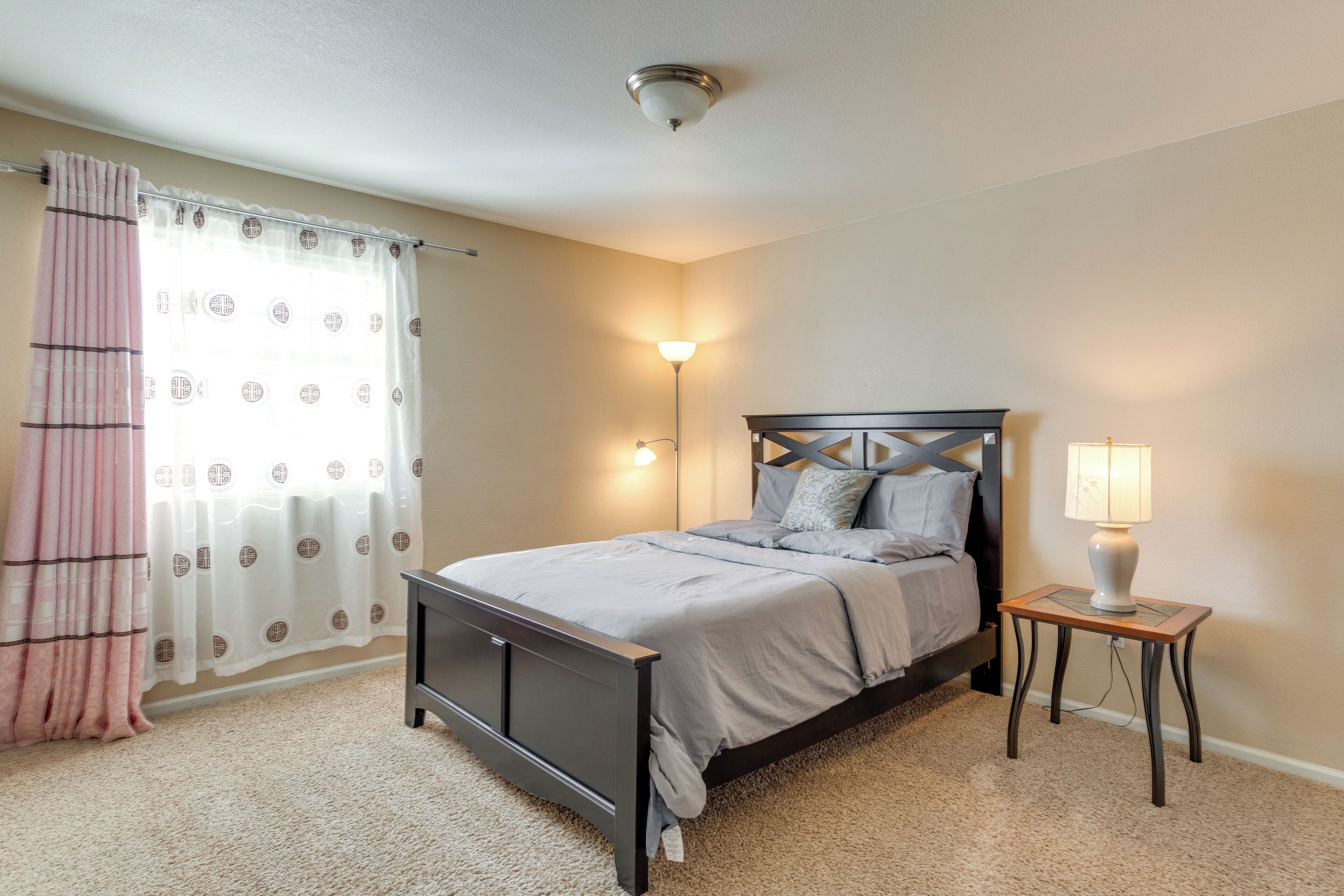 Une chambre à coucher avec un lit, une table de chevet avec une lampe, une fenêtre avec des rideaux et un luminaire fixé au mur.