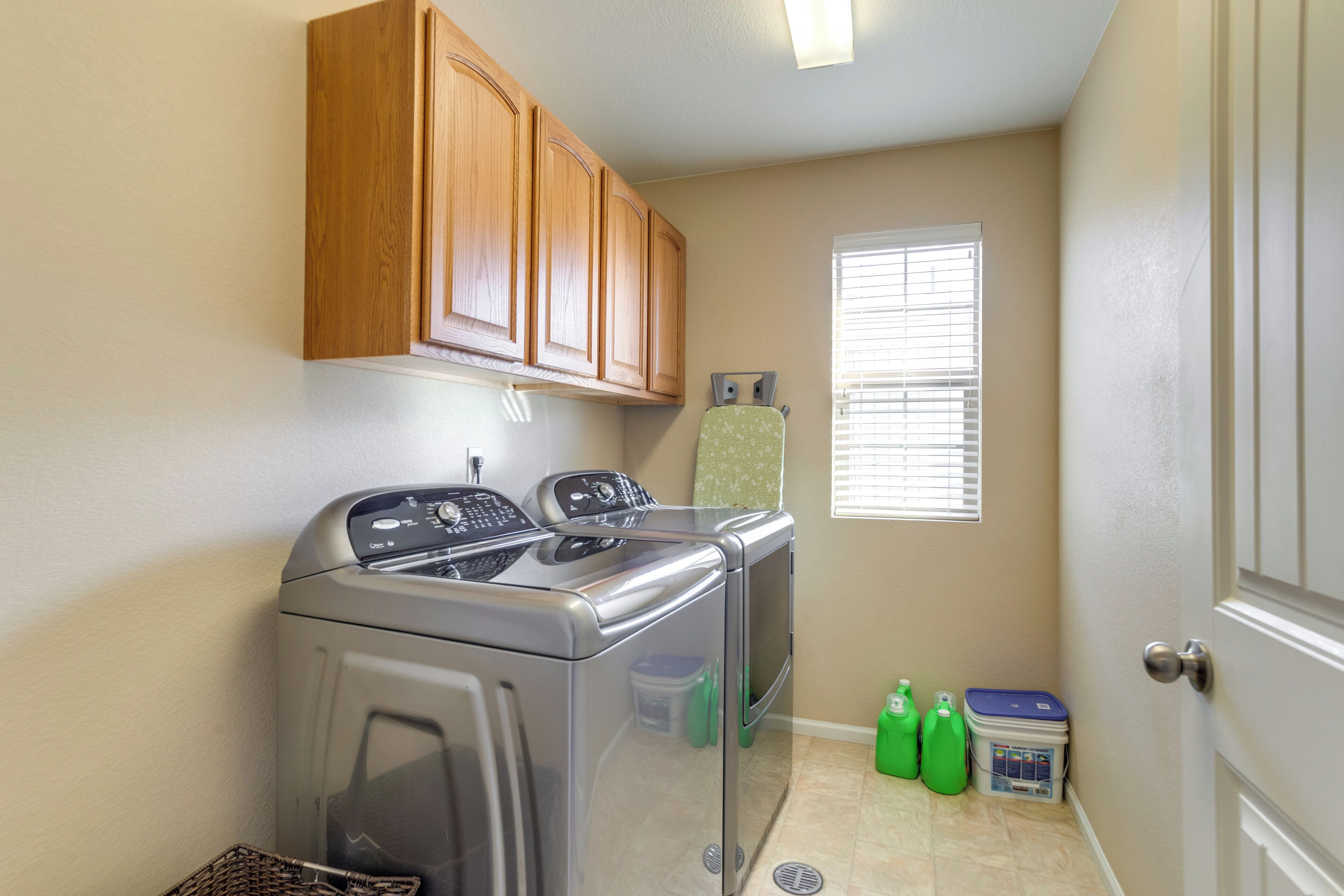 A laundry area with a washing machine and dryer, wooden cabinets, a window with blinds, and green cleaning supplies on the floor.