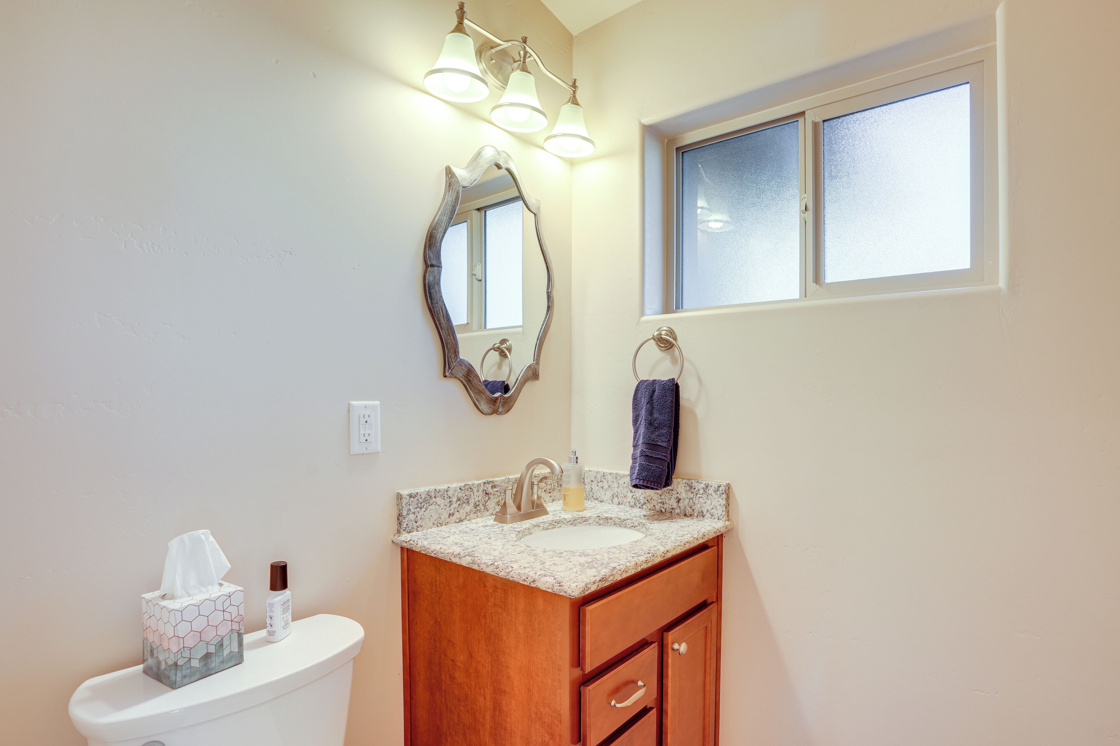 A bathroom with a toilet, sink, vanity, mirror, and window.