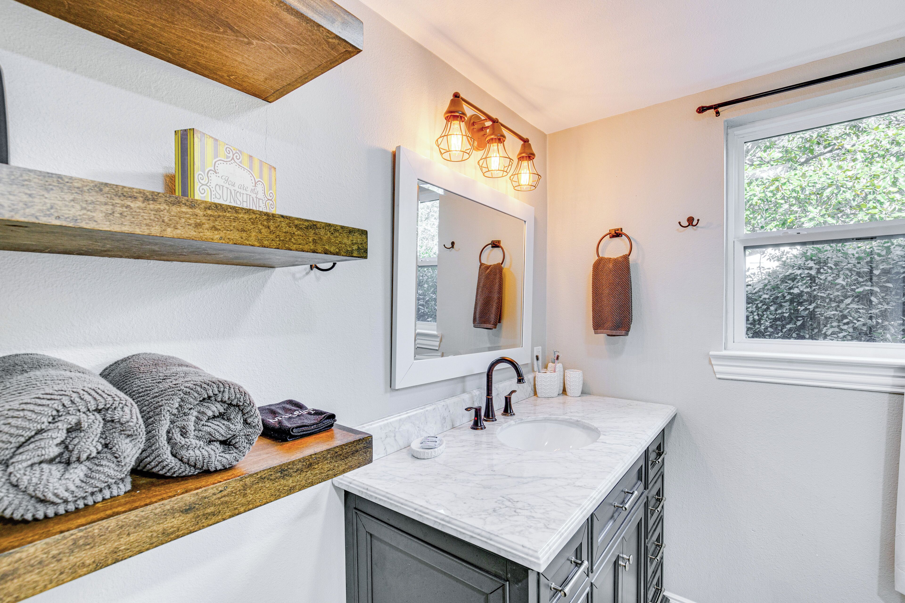 A bathroom with a marble sink, wooden shelves, a mirror, and a window with a view of trees.