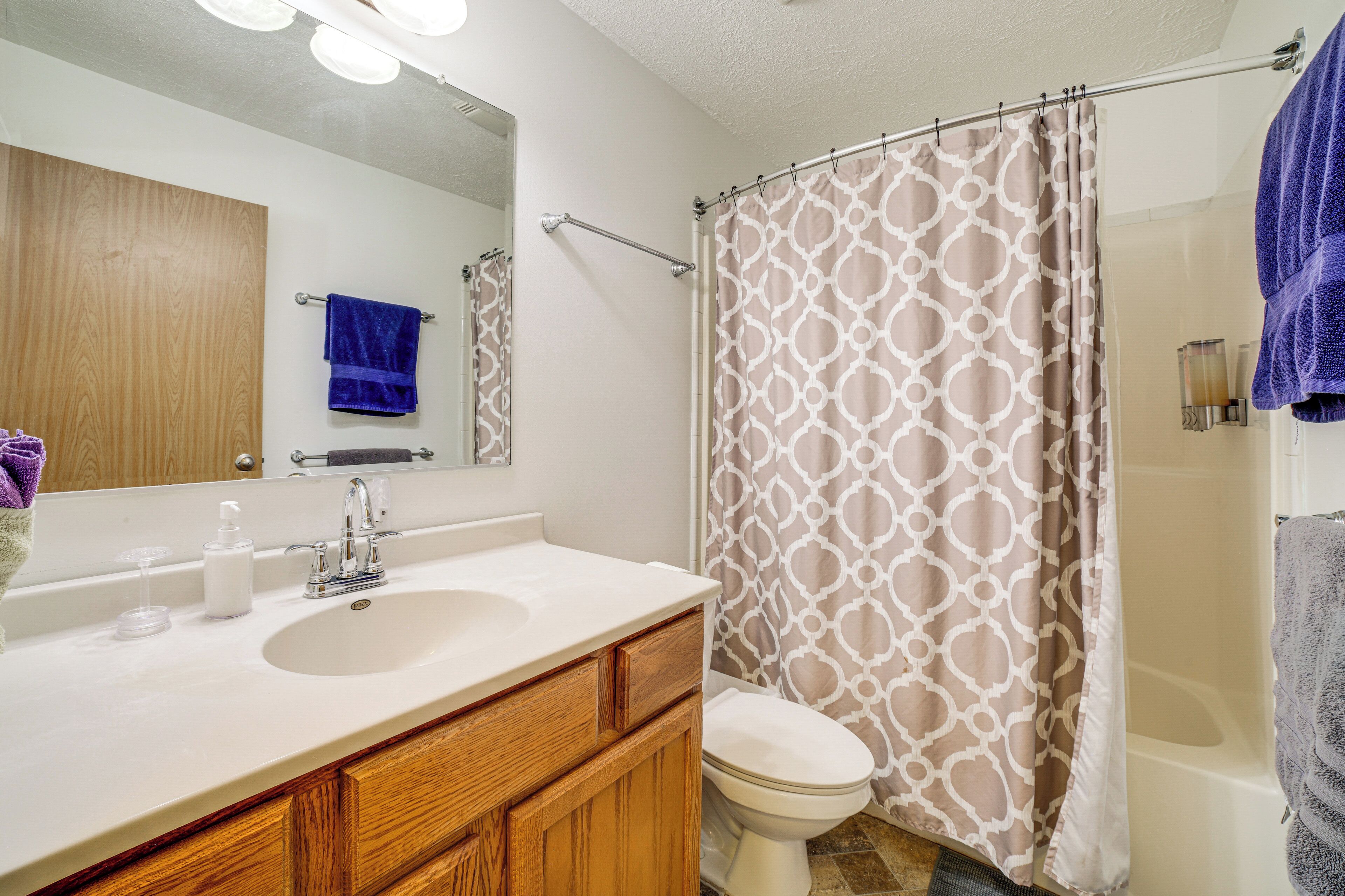Un baño con lavamanos blanco, tocador de madera, espejo, inodoro y ducha con cortina estampada.