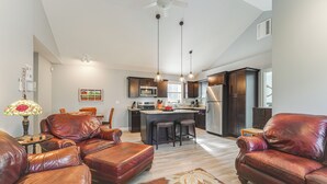 A spacious living room with leather sofas and a central island, a kitchen with dark cabinets and stainless steel appliances, and a ceiling fan. - Ski, Hike & Boat: Modern Bridgton Retreat (Bridgton)