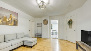 A spacious living room with a light-colored sectional sofa, a wooden floor, a flat-screen TV on a wooden stand, a cityscape painting, a round wall clock, and a door leading to a balcony. - NYC Area Apartment on Hudson River! (Hastings-On-Hudson)