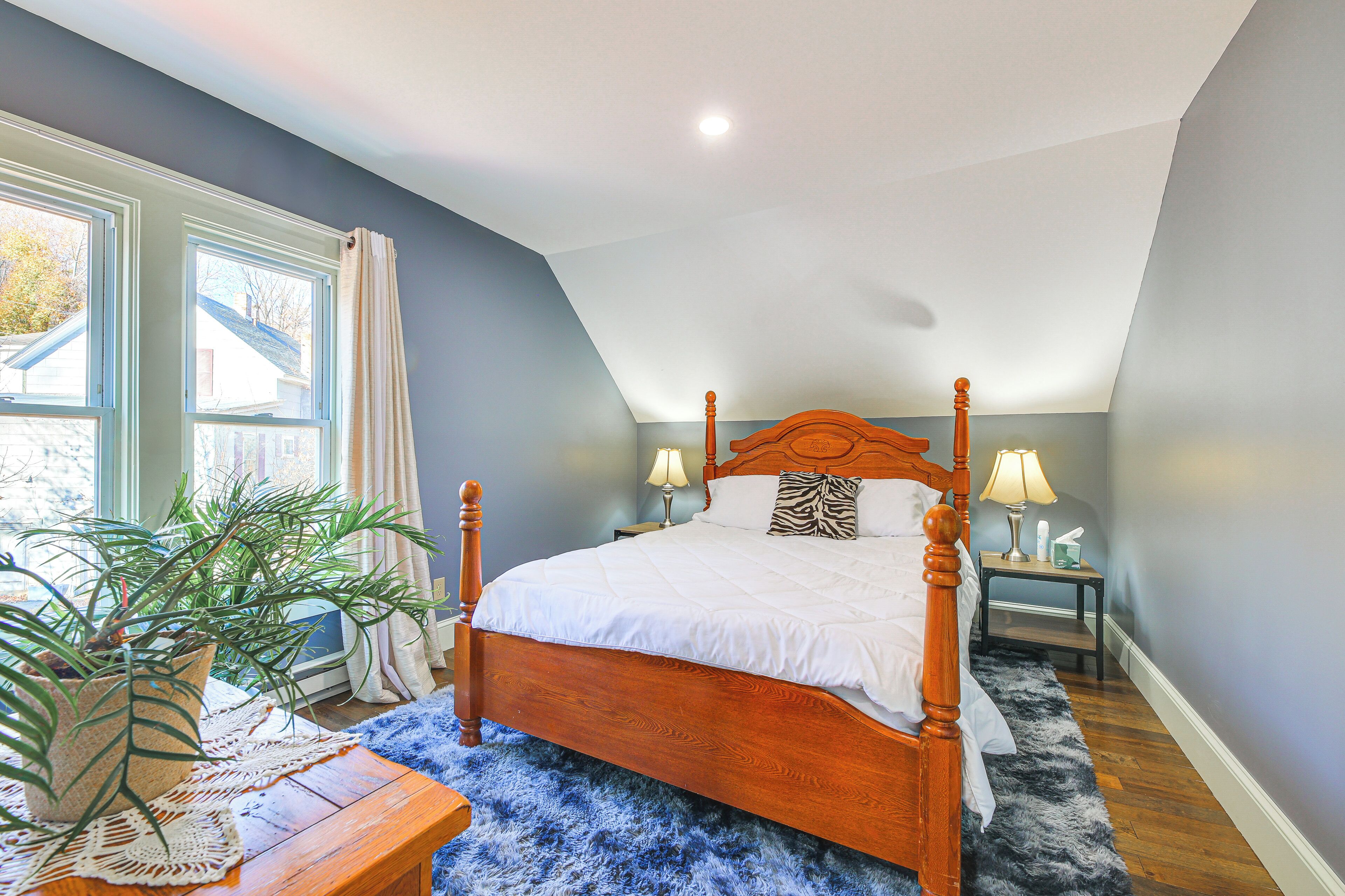 A bedroom with a wooden bed, a nightstand with a lamp, a potted plant, and a large window with curtains.