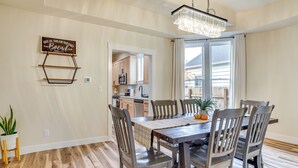 Un coin repas avec une table en bois, six chaises, un lustre et, au fond, une cuisine.