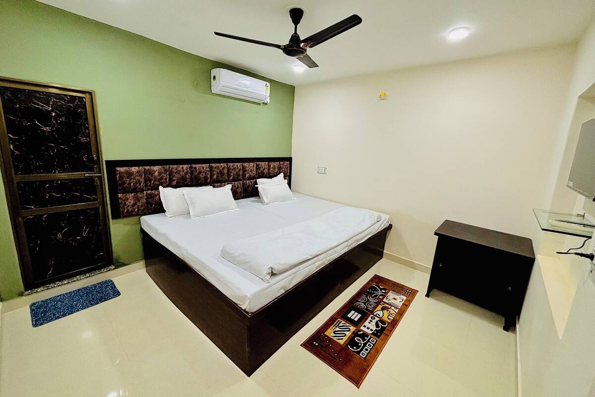 A hotel room with a bed, a ceiling fan, an air conditioning unit, a small desk, and a decorative rug.
