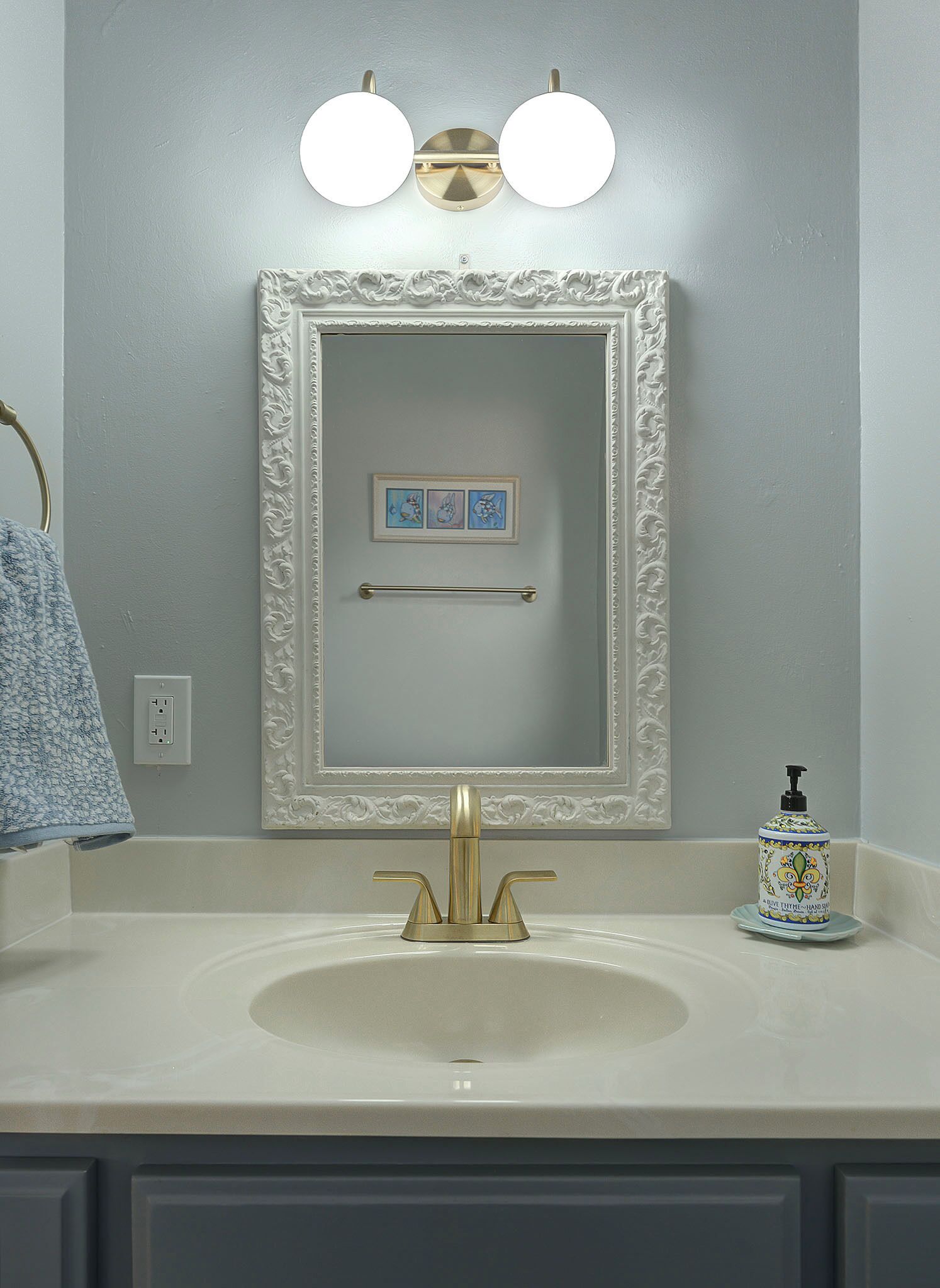 A bathroom with a white vanity, a gold faucet, a mirror with a decorative frame, and a soap dispenser on the counter.